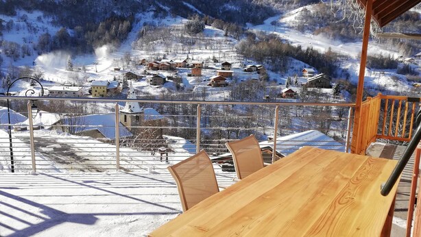 A wooden table and chairs on a balcony with a view of a snow-covered village and mountains.