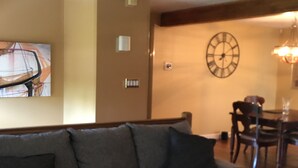 Un salon avec un canapé gris, une horloge au mur et un coin repas à l’arrière-plan.