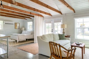 Una habitación con un sofá blanco, una silla de madera, una mesita, una cama con ropa de cama blanca y una ventana con cortinas.