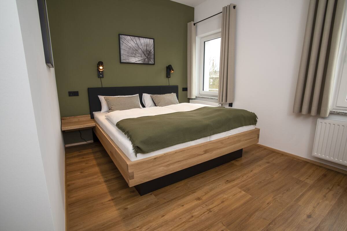 Une chambre moderne avec un lit en bois, un plaid vert et une grande fenêtre avec des rideaux.