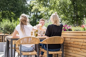 Three people sitting at an outdoor wooden table with wicker chairs, enjoying drinks and conversation.