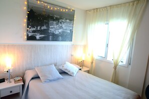 Quarto bem arrumado com cama, abajures, mesa de cabeceira, quadro na parede e janela com cortinas.