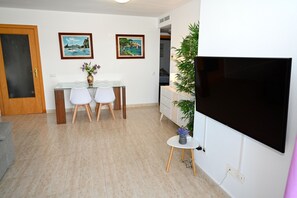 A modern living room with a flat-screen TV, a dining area with two chairs and a glass table, and two framed pictures on the wall.