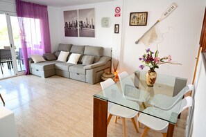 A modern living room with a glass dining table, white chairs, a grey sofa, and a cityscape painting on the wall.