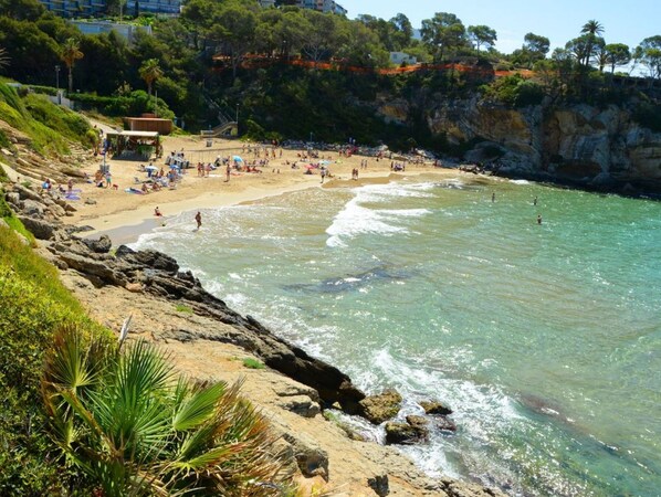 Una playa con aguas cristalinas, costa rocosa y algunos edificios al fondo.