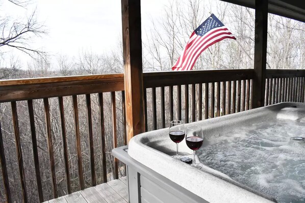 A hot tub on a wooden deck with two glasses of red wine, an American flag in the background, and leafless trees.