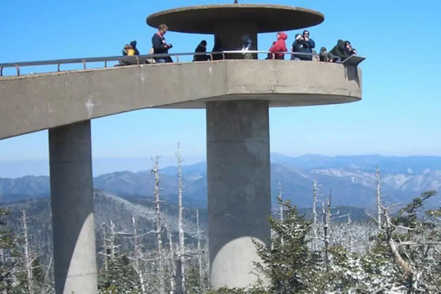 A concrete observation tower with a circular viewing platform, surrounded by snow-covered trees and mountains in the background.