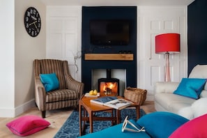 Una sala de estar con chimenea, televisor montado encima, un sillón a rayas, un sofá blanco y una lámpara de pie roja.