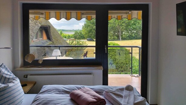 A bedroom with a large window overlooking a scenic landscape, a bed with pillows, a bedside table, and a radiator. - Pension Petersen (Bergen auf Ruegen)