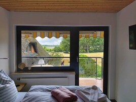 Un dormitorio con una ventana grande que da a un paisaje pintoresco, una cama con almohadas, una mesita de noche y un radiador.
