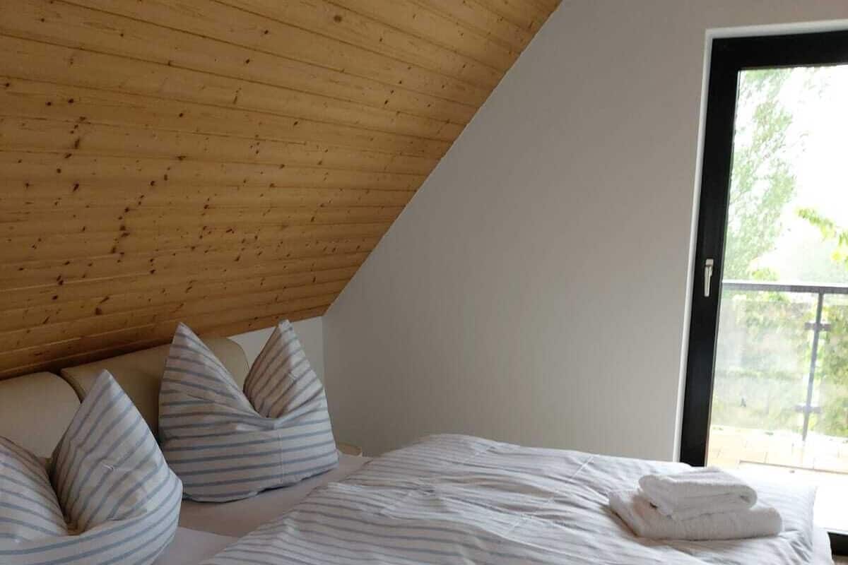Une chambre avec un plafond en pente, une grande fenêtre, un lit avec une literie à rayures et des oreillers.