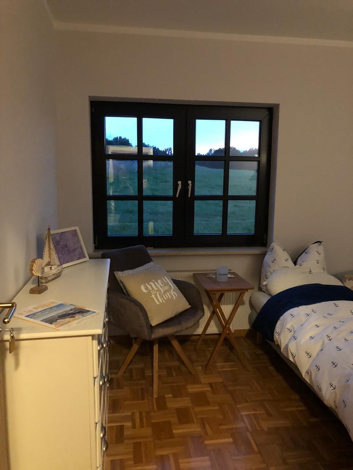 Una habitación pequeña con una cama, un escritorio y una silla. Se ve un ventanal con vistas a un campo.
