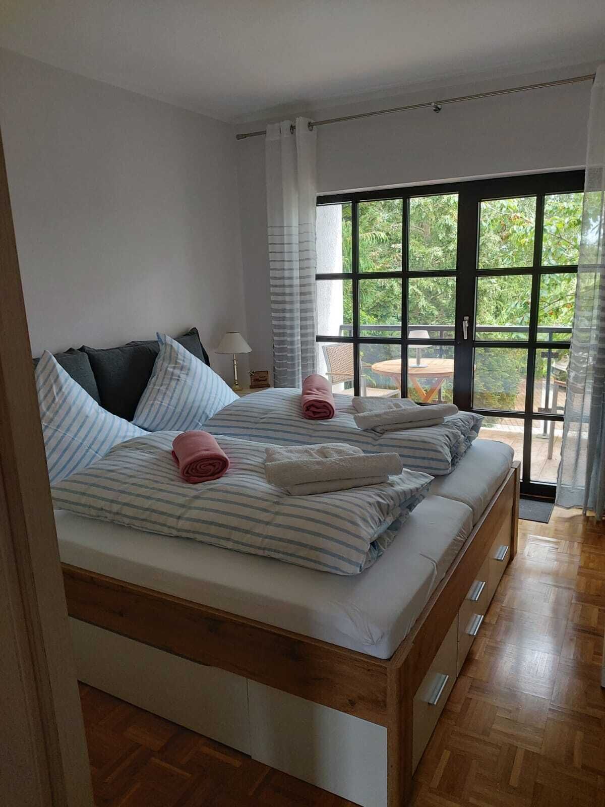 A double bed with striped bedding and rolled towels, a bedside table with a lamp, and a view of greenery through a large window.