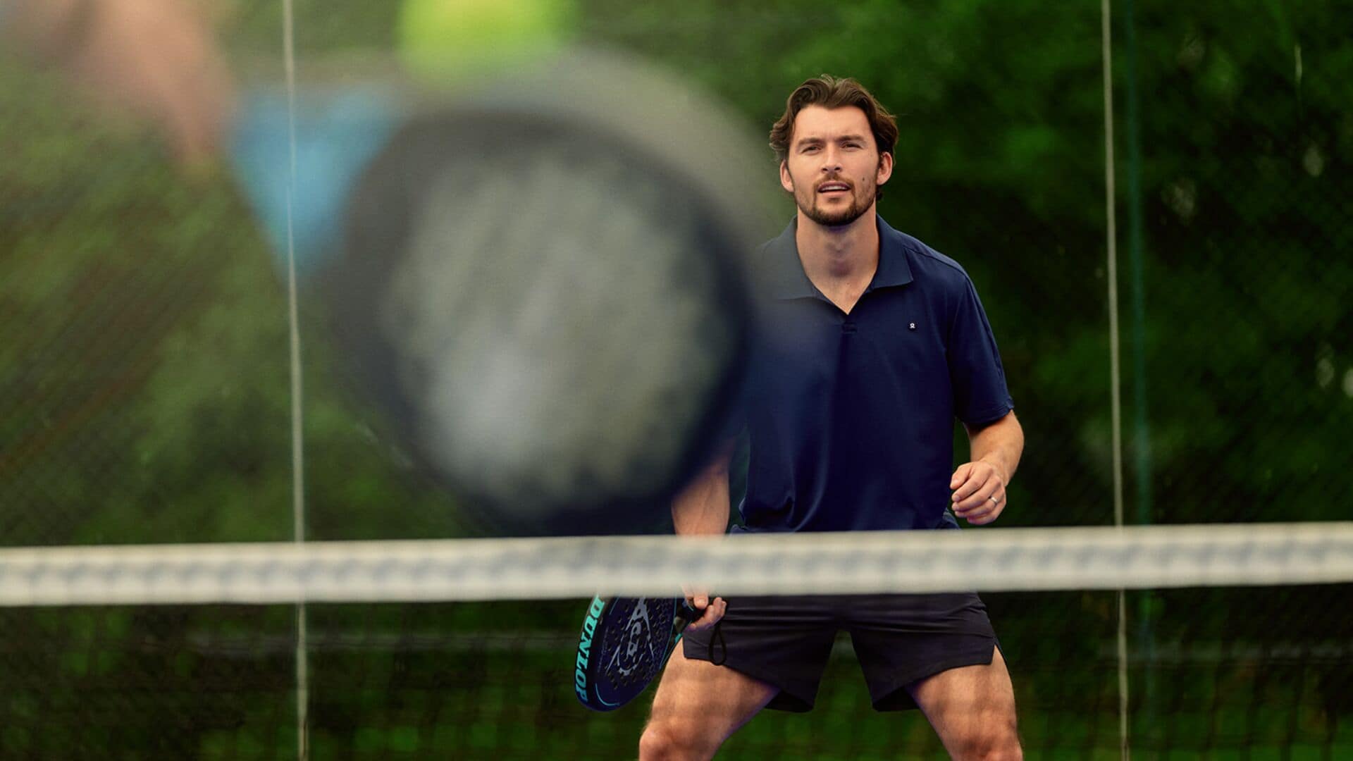 A tennis player in a dark polo shirt and shorts, holding a tennis racket, with a blurred tennis ball in motion.
