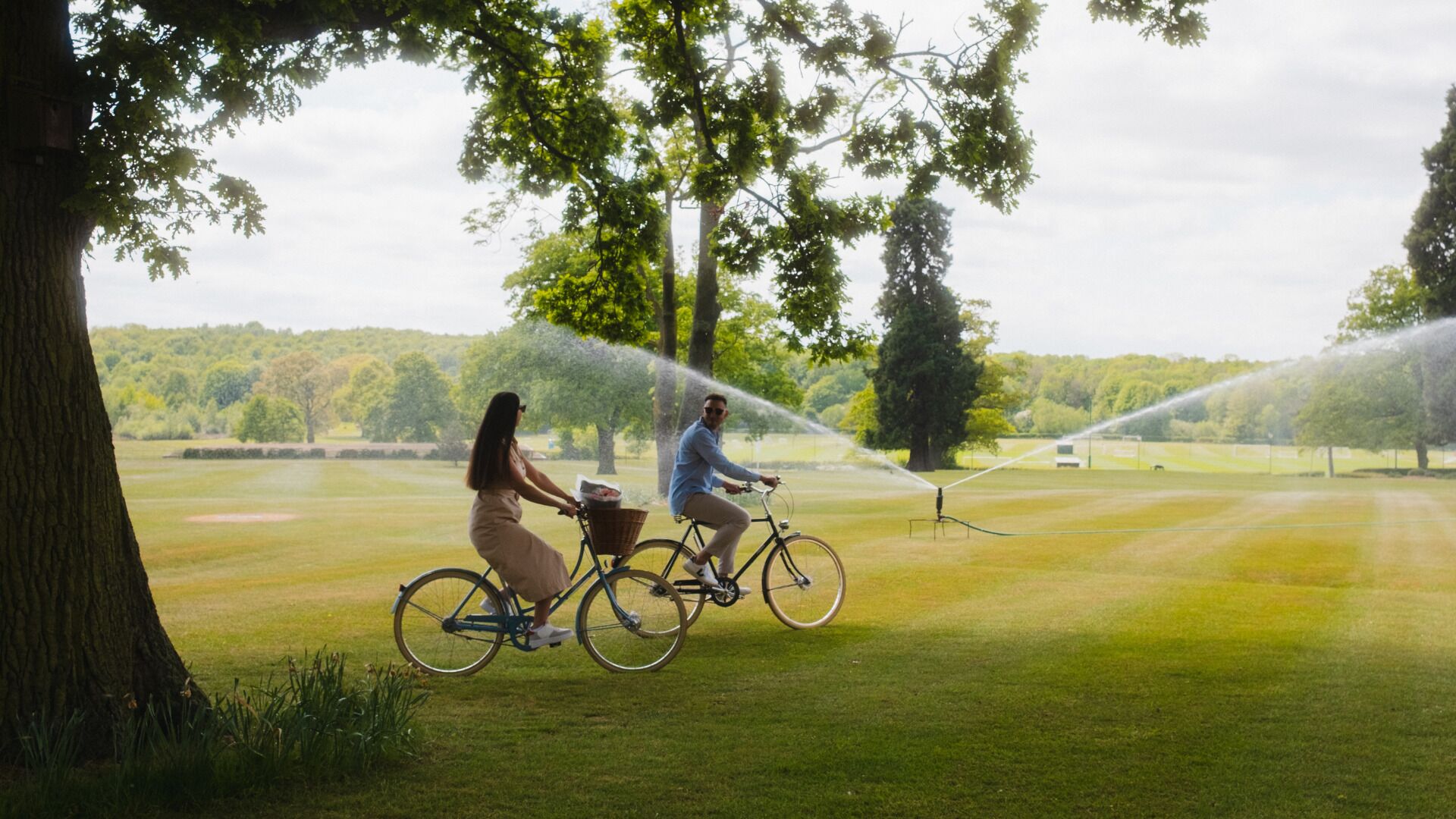 Una pareja pedalea en bicicleta sobre un campo cubierto de césped, con árboles y un sistema de riego al fondo.