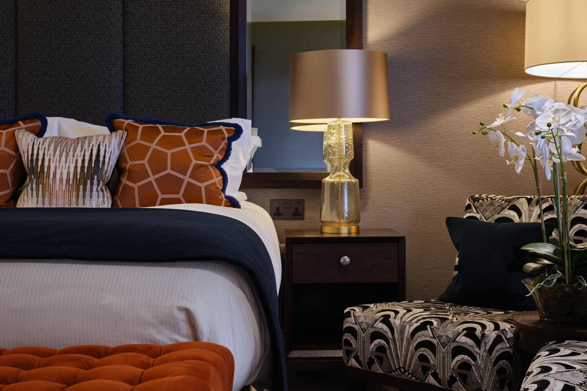 A neatly arranged hotel room with a bed, a bedside table with a lamp, a small table with a potted orchid, and a patterned ottoman.