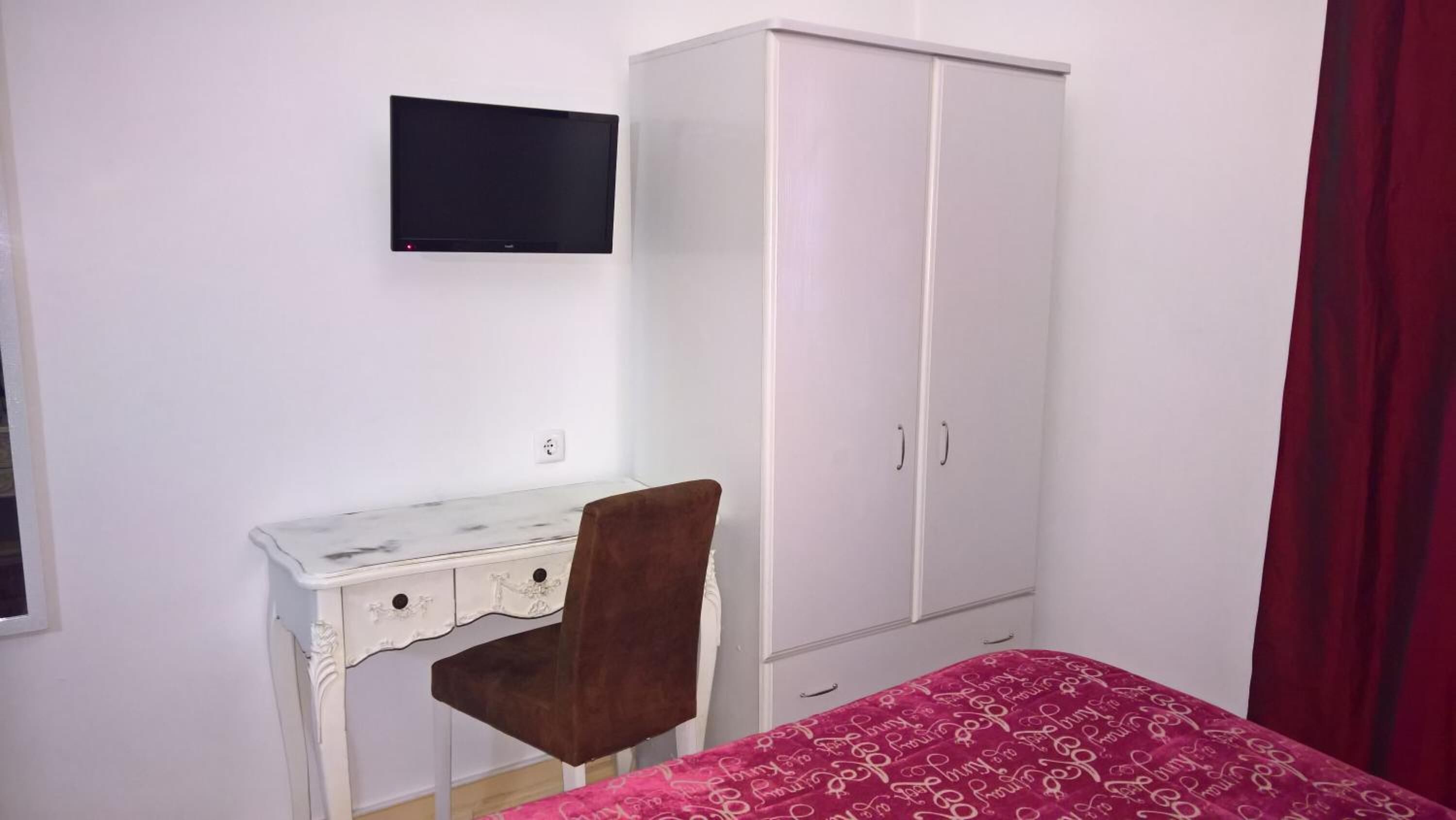 Una camera da letto con un armadio bianco, una scrivania con sedia, un letto con copripiumino fantasia rosa e una TV a schermo piatto montata a parete.