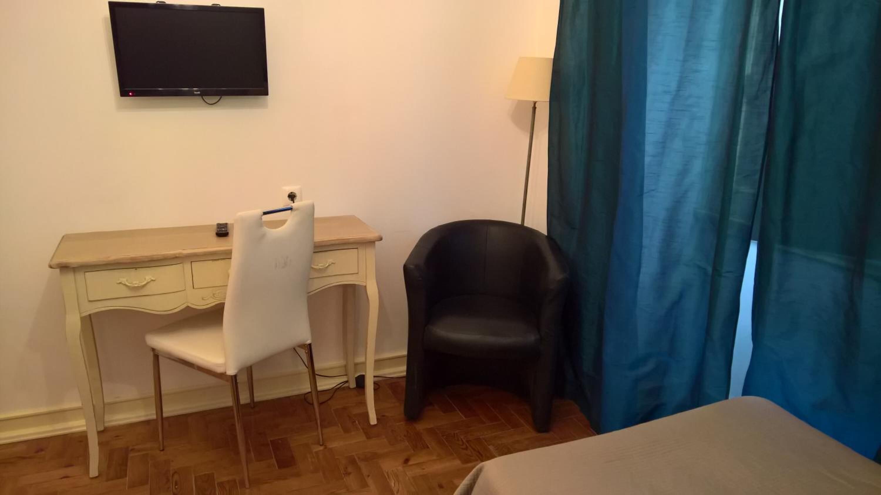 Une pièce avec un bureau en bois, une chaise blanche, une chaise noire, un lit, un téléviseur fixé au mur et une lampe.