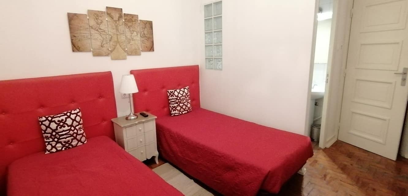 Quarto com duas camas vermelhas, uma mesinha de cabeceira com abajur e uma obra de arte na parede.