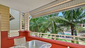 Un balcon avec une table et des chaises en verre, donnant sur un espace piscine agrémenté de palmiers et offrant une vue sur un ciel dégagé.