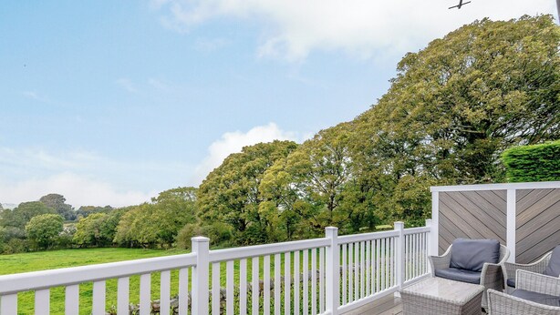 Une terrasse en bois avec des balustrades blanches, des meubles en osier et une vue sur un champ verdoyant et des arbres.