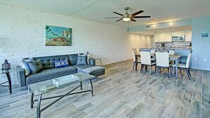 A living room with a sofa, coffee table, and dining area with chairs. - A Salty Day Getaway| Beach Boardwalk | Large Pool! (Port Aransas)