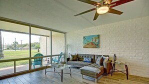 A spacious living area with a large window, a ceiling fan, a sofa, and a small table. - A Salty Day Getaway| Beach Boardwalk | Large Pool! (Port Aransas)