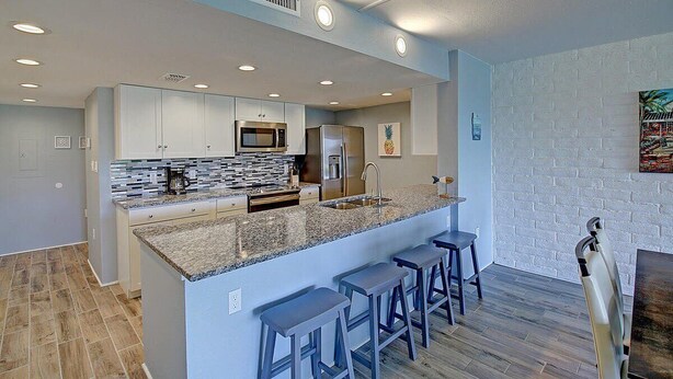 A modern kitchen with white cabinets, granite countertops, and stainless steel appliances. There are three bar stools and a white chair. - A Salty Day Getaway| Beach Boardwalk | Large Pool! (Port Aransas)