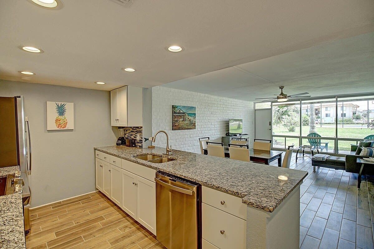 A modern kitchen with granite countertops, white cabinetry, and a dishwasher. There is a dining area with a table and chairs, and a large window offering a view of the outdoors.