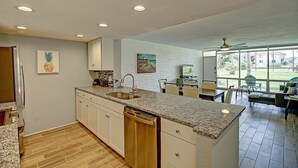 A modern kitchen with granite countertops, white cabinetry, and a dishwasher. There is a dining area with a table and chairs, and a large window offering a view of the outdoors. - A Salty Day Getaway| Beach Boardwalk | Large Pool! (Port Aransas)