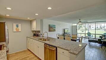 A modern kitchen with granite countertops, white cabinetry, and a dishwasher. There is a dining area with a table and chairs, and a large window offering a view of the outdoors.