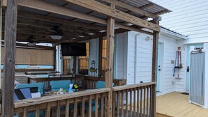 Une terrasse extérieure couverte avec des rampes en bois, un coin salon avec des coussins bleus, et un espace cuisine à l’arrière-plan.