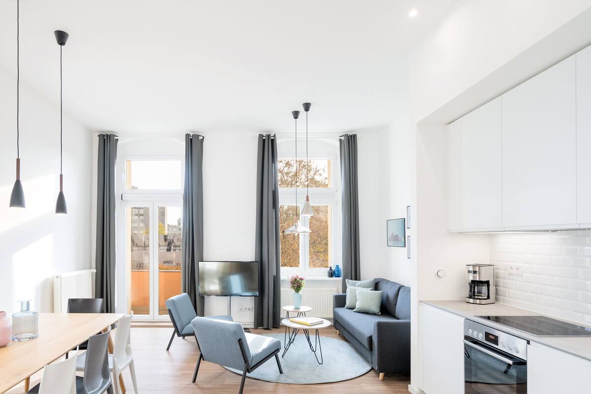 A modern living room with a dining area, a kitchen, and a large window with curtains.