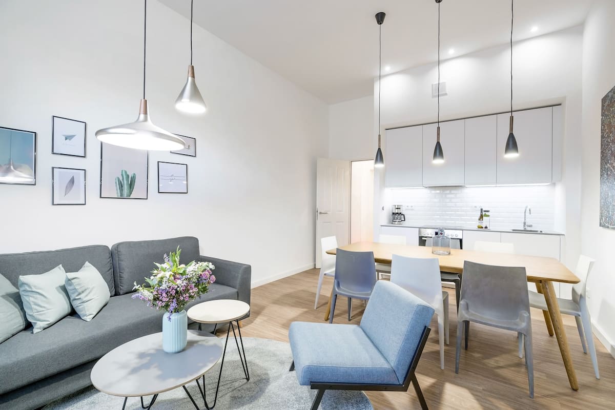 A modern living room with a grey sofa, a blue armchair, a round coffee table, and a small white table with a vase of flowers. There is a kitchen area with a wooden table, chairs, and a white cabinet.