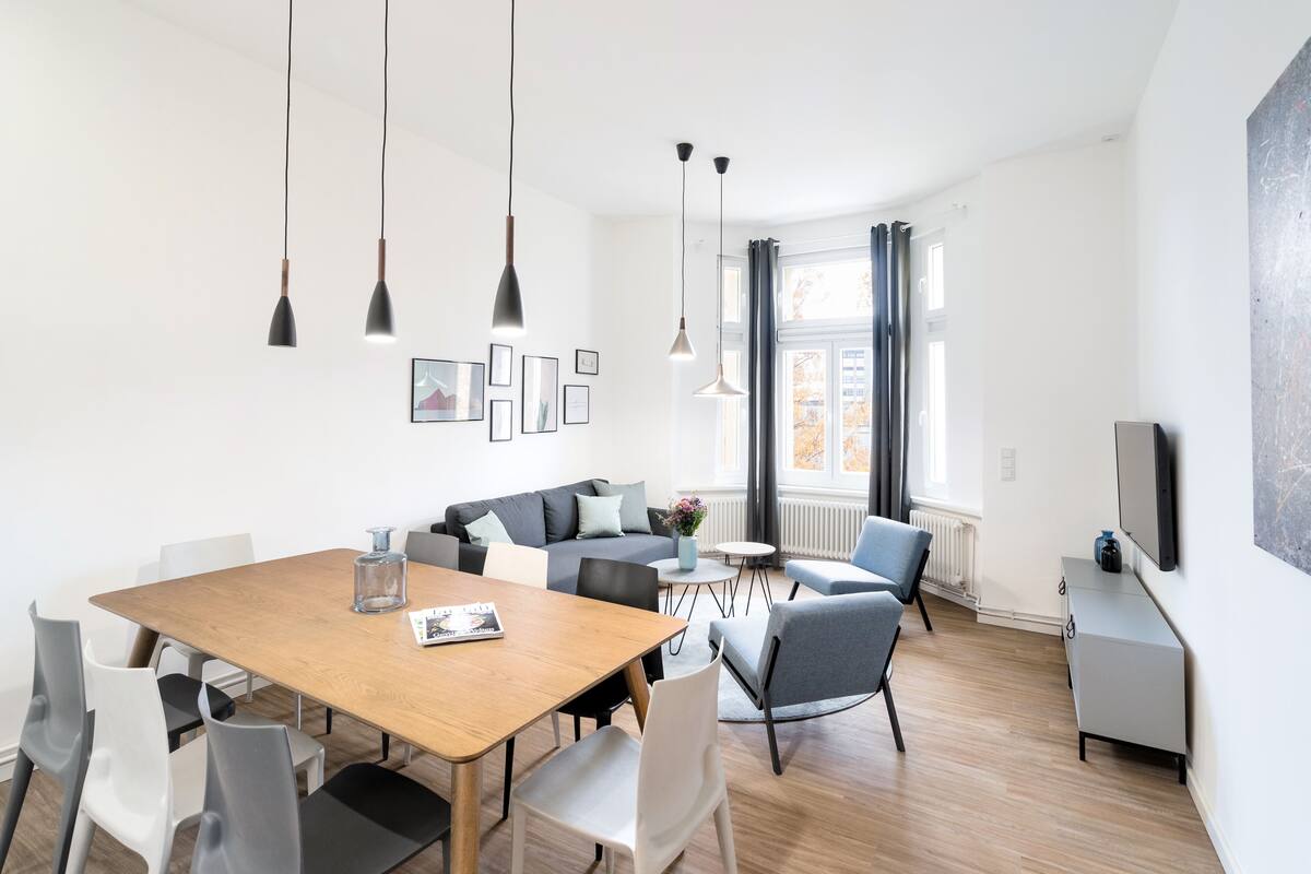 A modern living room with a wooden dining table, grey and white chairs, a grey sofa, and a grey armchair. There are pendant lights, a coffee table, and a TV mounted on the wall.