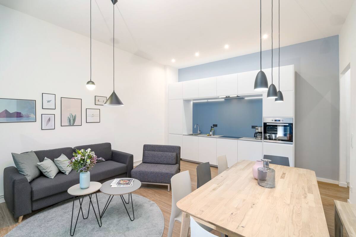 A modern living room with a sofa, a coffee table, and a dining table. There is a kitchen area with cabinets and a sink.