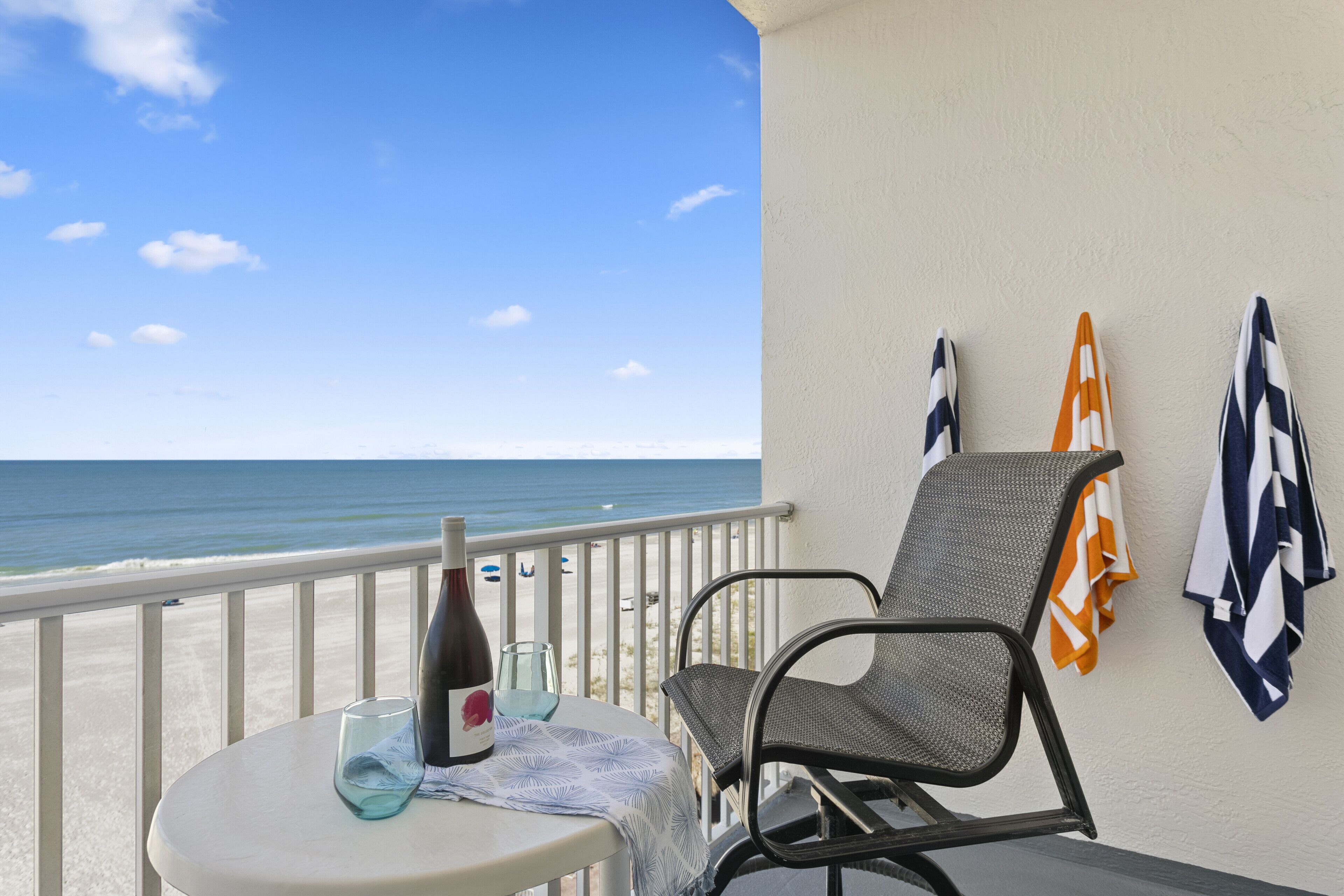 A balcony with a table, a chair, a bottle of wine, and a glass. There are towels hanging on the wall. The view includes the ocean and a clear sky.