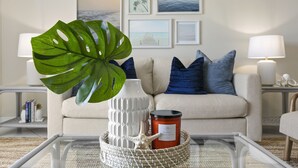 Sala de estar con una mesa de centro de vidrio, un jarrón blanco, una vela roja y una planta verde grande. Hay un sofá color beige con almohadas azules y blancas, y una lámpara blanca sobre una mesita de noche.