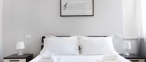 Uma cama bem arrumada com lençóis e almofadas brancas, uma partitura emoldurada na parede e duas mesas de cabeceira com abajures.