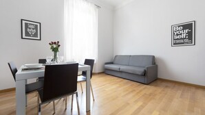 A modern living room with a dining area. There is a grey sofa, a dining table with chairs, and a vase with flowers. Two framed pictures are on the walls. - UR-NEST Marconi 2, Gorgeous and spacious one bedroom apartment (Bologna)