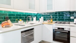A modern kitchen with white cabinets, a green tiled backsplash, stainless steel appliances, and a bag of oranges labeled "BOLLO".