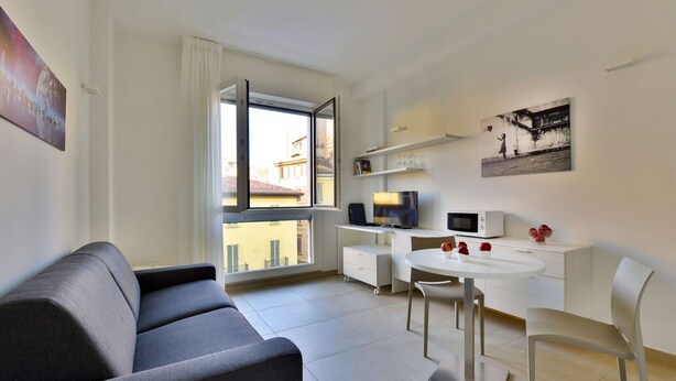 Un salon moderne avec un canapé gris, une table à manger avec des chaises, un bureau blanc avec un ordinateur et un micro-ondes, et une grande fenêtre donnant sur des immeubles.