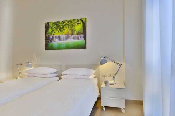 Habitación de hotel con dos camas, una mesita de noche con una lámpara, un cuadro de una cascada y una cortina blanca.