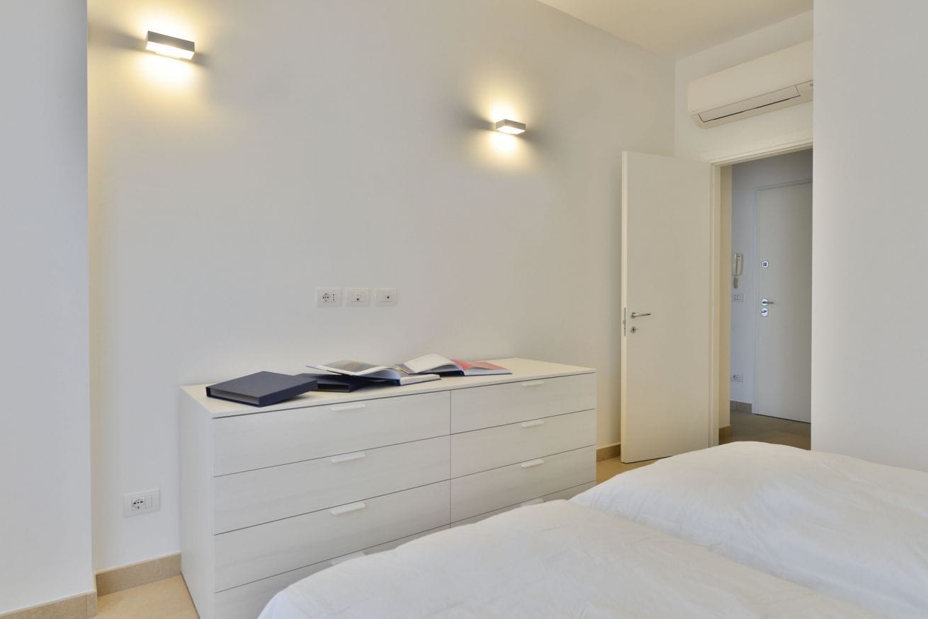 A modern bedroom with a white bed, a bedside table with books, and two wall-mounted lights.