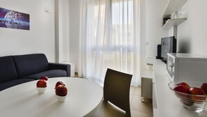 Un salon moderne avec une table de salle à manger ronde, un canapé foncé, un téléviseur et un coin cuisine équipé d’un four à micro-ondes.
