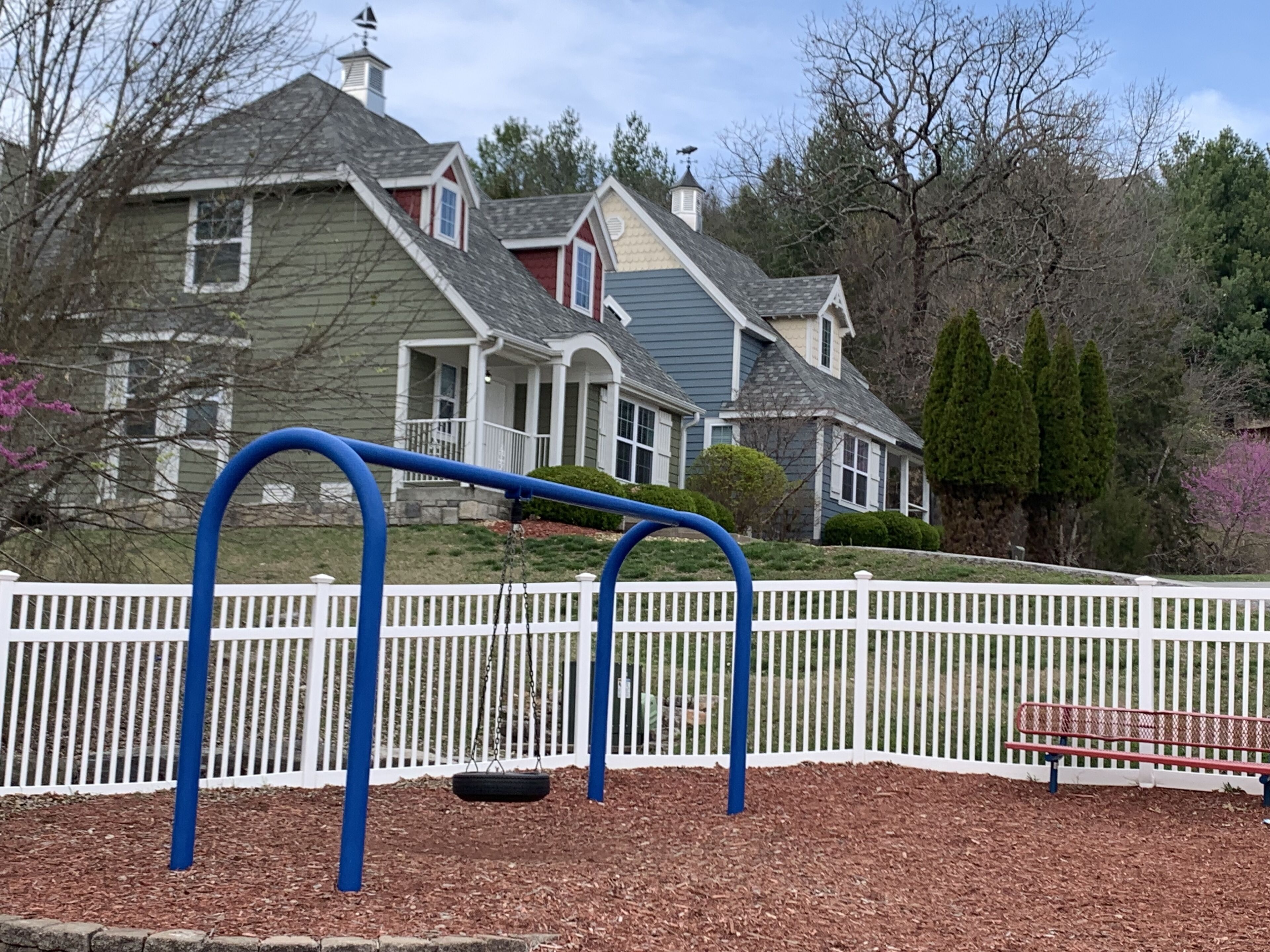 A playground with swings and a bench, surrounded by a white picket fence, with houses in the background.