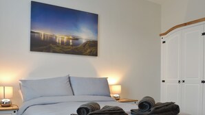 A neatly made bed with a blue and white checkered bedspread, two pillows, and a pair of folded towels on a bedside table. A painting of a night scene with a bridge is hung on the wall above the bed.