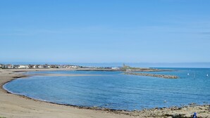 A coastal scene with a sandy beach, calm blue waters, and a distant shoreline with buildings and a lighthouse.