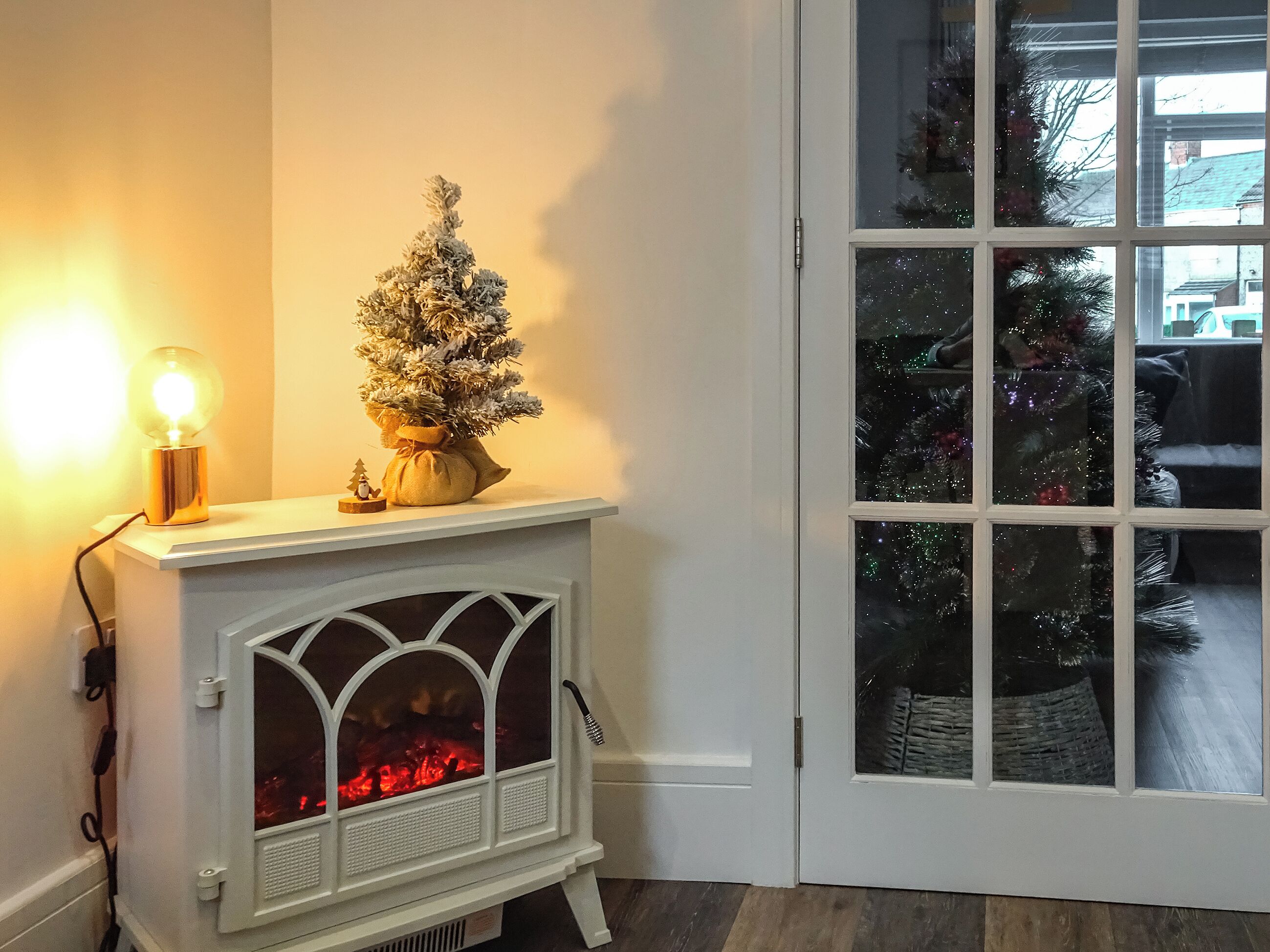 Een gezellige kamer met een witte open haard, een kleine kerstboom en een brandende lamp.