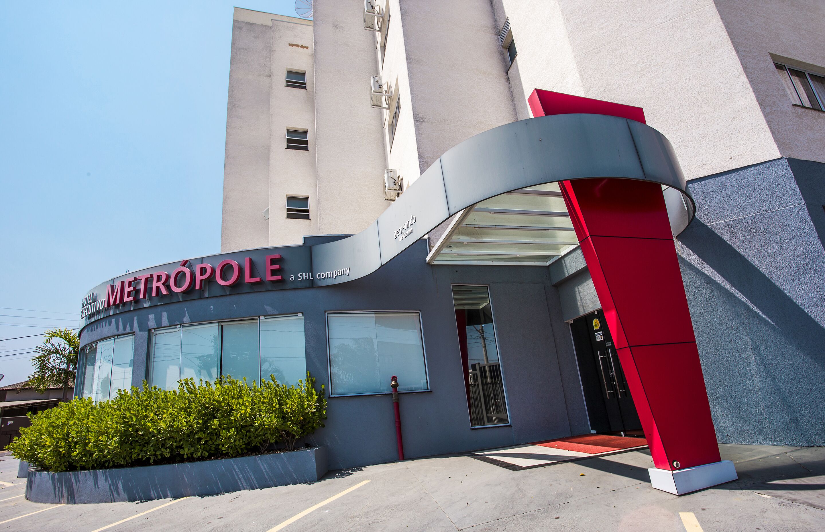 Entrada de hotel moderna com fachada curva em vermelho e cinza, grandes janelas e um tapete vermelho que leva à porta.